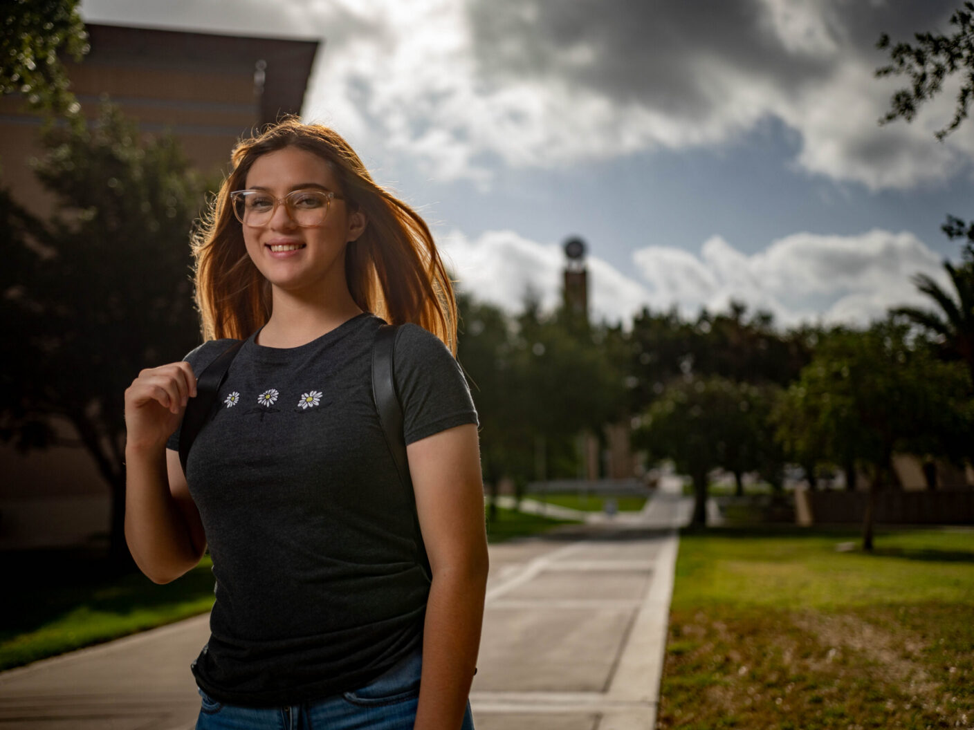 A young person with long hair and glasses smiles while walking outdoors on a paved path, wearing a dark t-shirt with small flower designs and carrying a backpack.