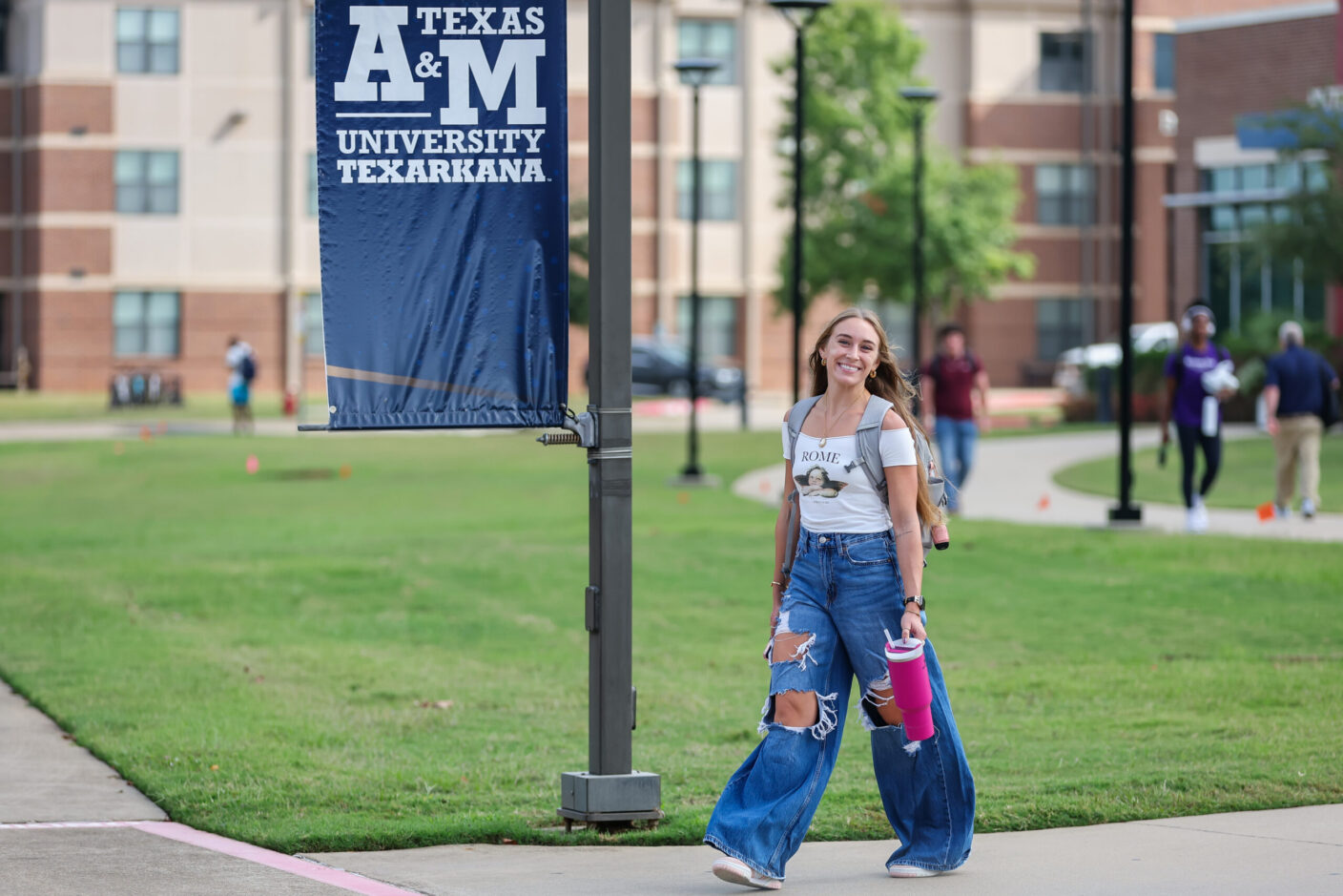 A person walks across a campus pathway near a Texas A&M University-Texarkana banner, wearing ripped jeans and carrying a pink cup.