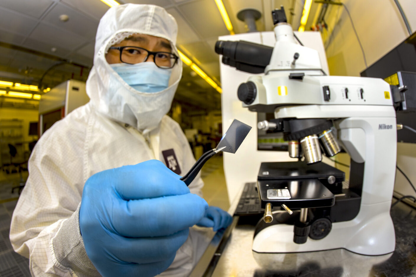 A scientist in a lab coat and mask holds a small sample with tweezers near a Nikon microscope.