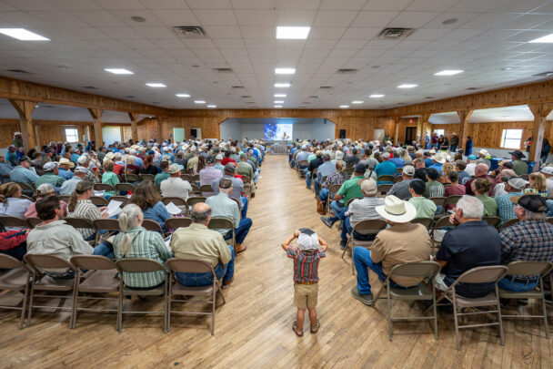 A large auditorium with a speaker down the center and a slideshow in the background.